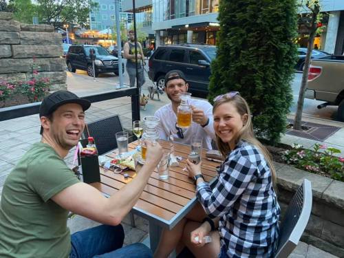 Three friends smiling and toasting with drinks at an outdoor table, surrounded by greenery and a lively street scene.