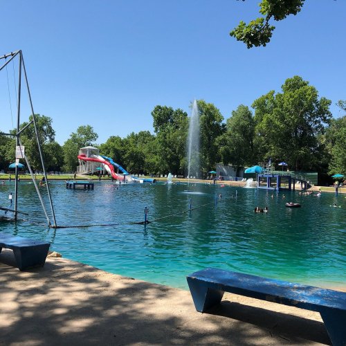 A sunny outdoor pool area with a water slide, trees, and people swimming and relaxing by the water.