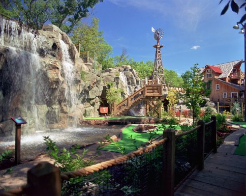 A scenic outdoor area featuring a waterfall, windmill, and mini-golf surrounded by lush greenery and a blue sky.