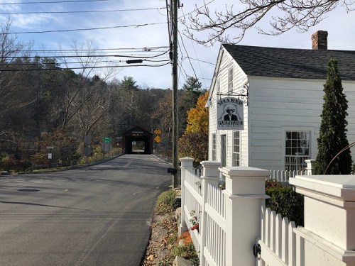 A white house with a sign near a road leading to a covered bridge, surrounded by trees and autumn foliage.