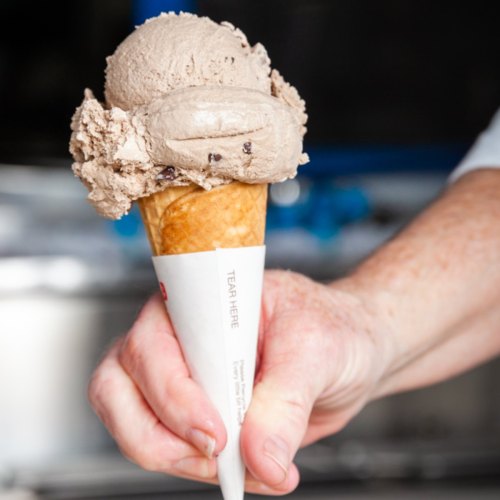 A hand holding a cone with a scoop of chocolate ice cream on top, set against a blurred background.