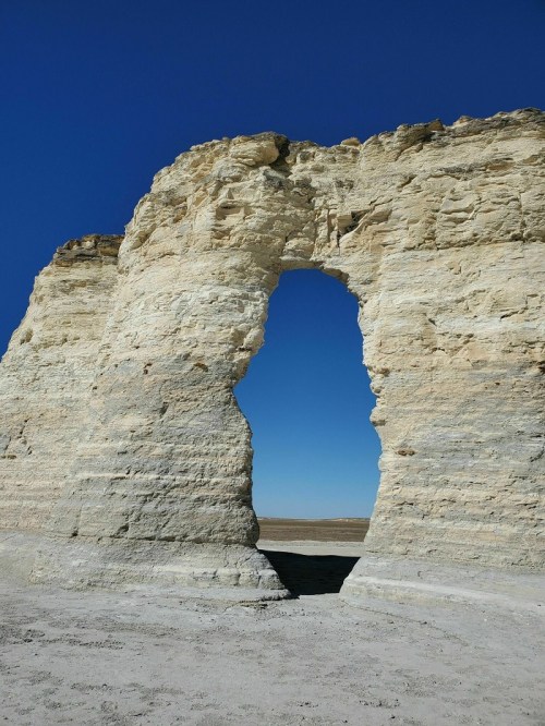 A large rock formation with a natural arch against a clear blue sky, set in a barren landscape.