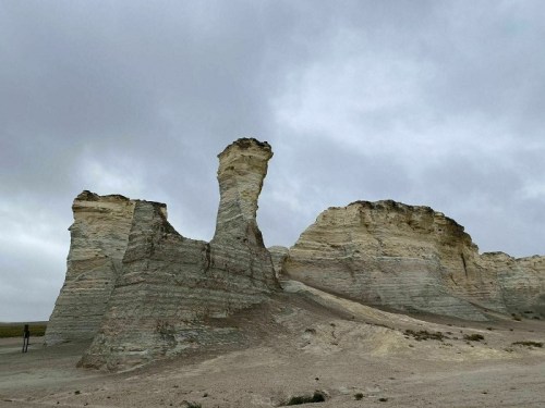 A rocky formation with a tall, slender spire stands against a cloudy sky in a barren landscape.