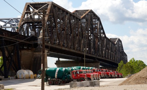 A large metal bridge spans over a construction site with red cement trucks parked underneath.