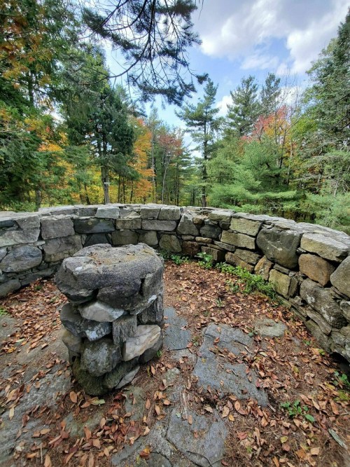 A circular stone structure surrounded by trees, with autumn foliage and a cloudy sky in the background.