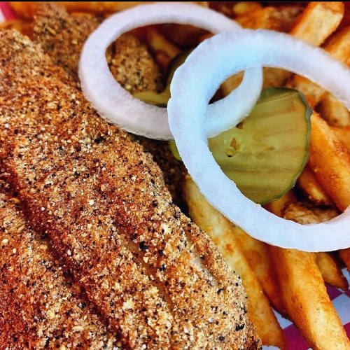 A close-up of seasoned fish fillet with onion rings and pickles, served alongside crispy French fries.
