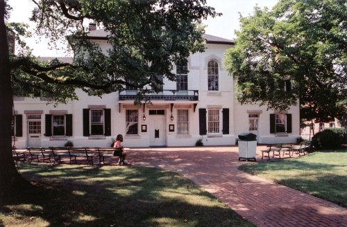 A white historic building with green shutters, surrounded by trees and benches, on a sunny day.