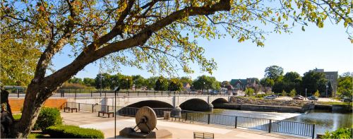 A scenic view of a river with a bridge, surrounded by trees and park benches on a sunny day.