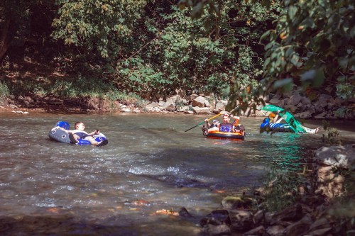 Two people float on inflatable tubes in a river, surrounded by trees and rocky banks on a sunny day.