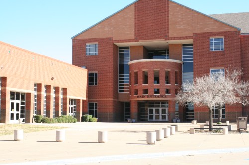 A modern brick building with large windows and a prominent main entrance, surrounded by a paved area and sparse landscaping.