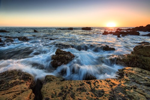 Sunset over the ocean, with waves crashing against rocky shorelines and a warm glow in the sky.