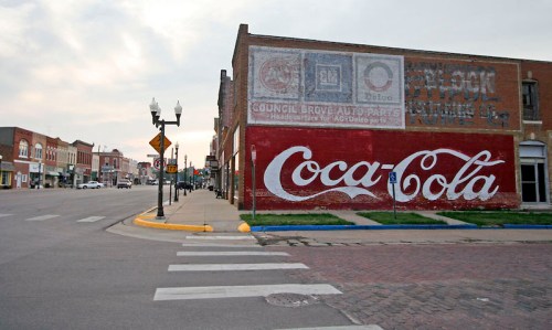 Faded Coca-Cola mural on a brick wall in a small town, with a street and buildings in the background.