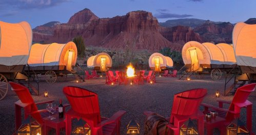 A cozy campsite with glowing wagons, red chairs, and a campfire, set against a stunning mountain backdrop at dusk.