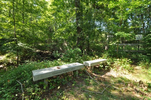 Two stone benches in a lush green forest, with a plaque reading "Cranston Foundry 1812" nearby.