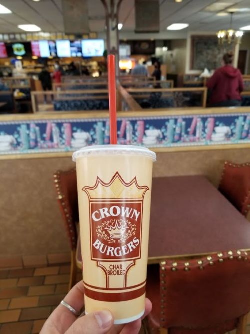 A hand holding a drink cup from Crown Burgers, with a restaurant interior visible in the background.