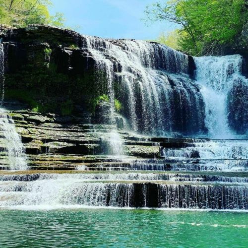 A serene waterfall cascading over rocky ledges into a clear blue pool, surrounded by lush green trees.
