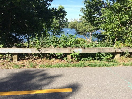 A view of a path beside a wooden railing, with greenery and a river visible in the background under a clear blue sky.