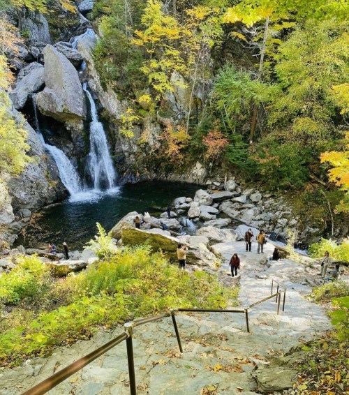A scenic waterfall surrounded by autumn foliage, with visitors exploring the rocky area and a staircase leading down.