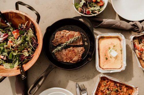 A table spread with a skillet of steak, a salad bowl, and various side dishes, including cornbread and baked beans.