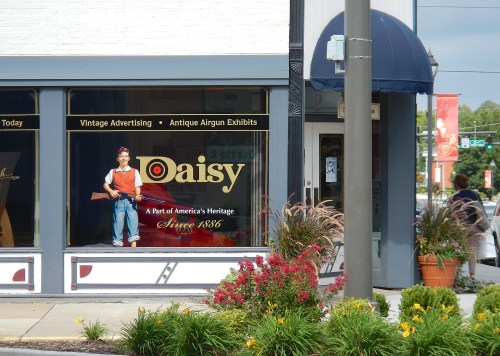 A storefront window displays a vintage advertisement for Daisy, featuring a child with an airgun and colorful flowers outside.