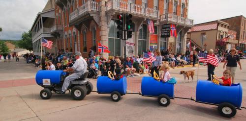 A parade scene with people watching as a man drives a small tractor pulling blue barrels, decorated with flags.