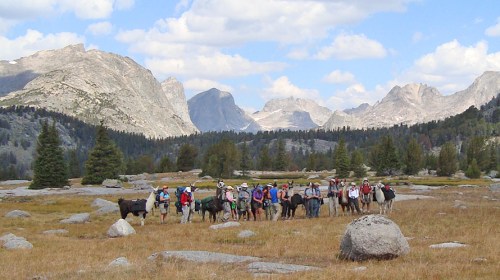 A group of hikers with pack animals in a mountainous landscape, surrounded by trees and rocky peaks under a blue sky.