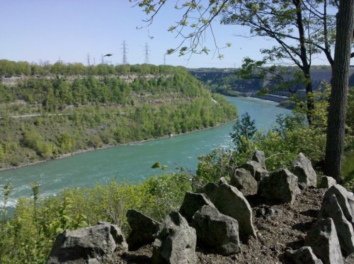 A scenic view of a river winding through lush green hills, framed by rocks and trees under a clear blue sky.