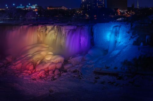 Niagara Falls illuminated at night with vibrant colors, surrounded by ice and snow.