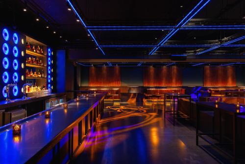 A modern bar interior with blue lighting, a long counter, shelves of bottles, and candlelit tables.