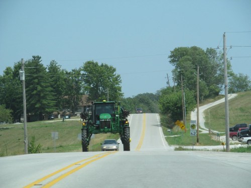 A green tractor drives down a rural road, with trees and a hill in the background under a clear blue sky.