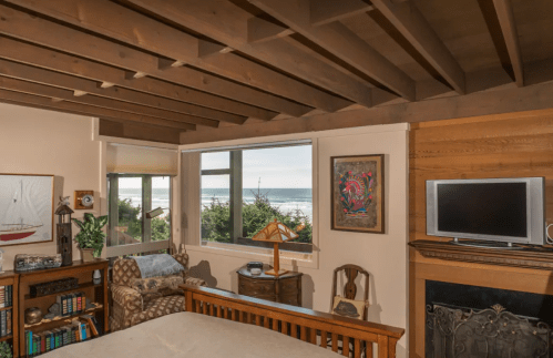 Cozy bedroom with wooden beams, a view of the ocean, and a fireplace, featuring a chair and decorative artwork.