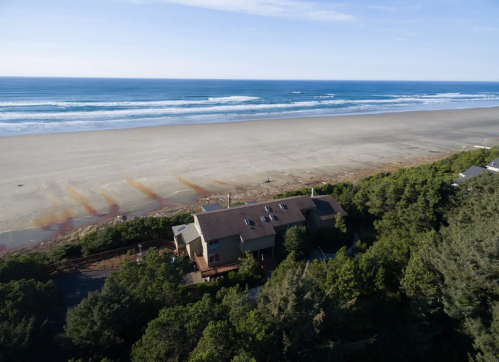 Aerial view of a beach house surrounded by trees, with a wide sandy beach and ocean waves in the background.