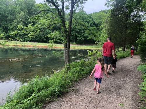 A man and two children walk along a path by a serene pond, surrounded by lush greenery.