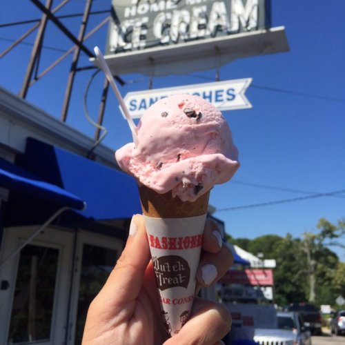 A hand holding a cone of pink ice cream with a sign for "Homemade Ice Cream" in the background on a sunny day.