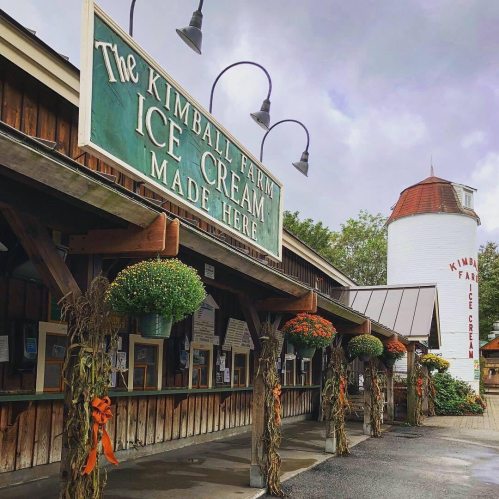 Sign for Kimball Farm Ice Cream with a rustic building and decorative plants, under a cloudy sky.