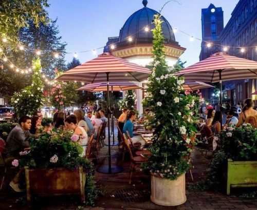 A lively outdoor restaurant scene at dusk, with diners under striped umbrellas surrounded by greenery and string lights.