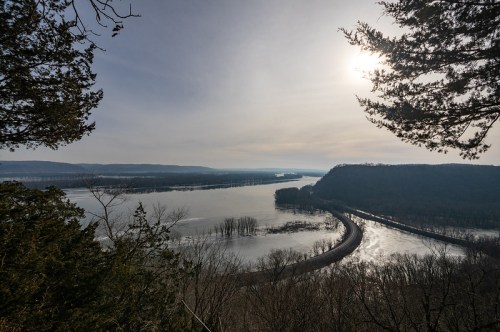 A scenic view of a winding river surrounded by trees, with a hazy sky and sun shining above.