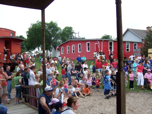 A large crowd of people, including children and families, gathered outdoors near a red building and train car.
