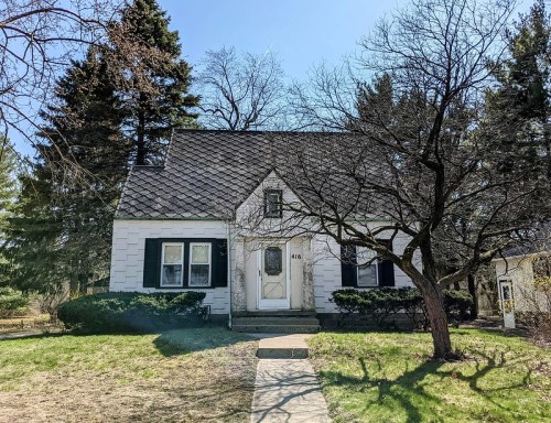 A charming white house with a steep roof, black shutters, and a front yard, surrounded by trees on a sunny day.