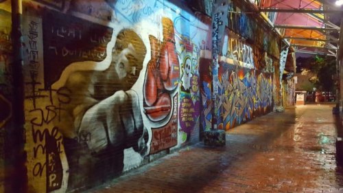 Colorful street art covers a brick wall in a dimly lit alley, featuring a boxing figure and vibrant graffiti.