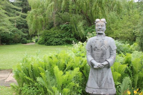 A stone statue of a warrior stands among lush green ferns in a serene garden setting.