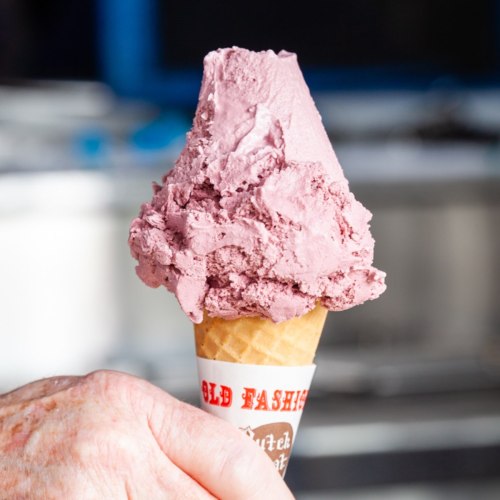 A hand holding a cone topped with a large scoop of purple ice cream against a blurred background.