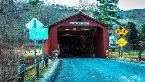 A red covered bridge with a wreath, leading into a wooded area, with road signs indicating town lines and a one-lane bridge.