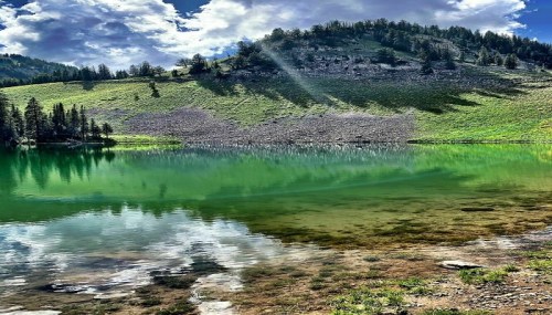 A serene lake reflecting green hills and a cloudy sky, surrounded by trees and rocky terrain.