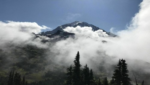 A majestic mountain peak partially covered by clouds, surrounded by lush greenery and tall evergreen trees.