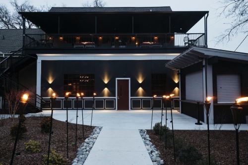 A modern building with a dark exterior, lit by torches along a pathway, and a balcony overlooking the entrance.