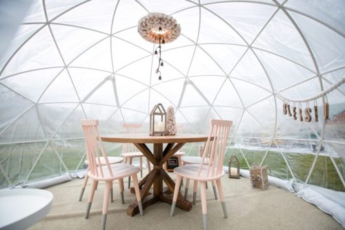 A cozy, transparent igloo-style dining space with a wooden table, pink chairs, and decorative lanterns.