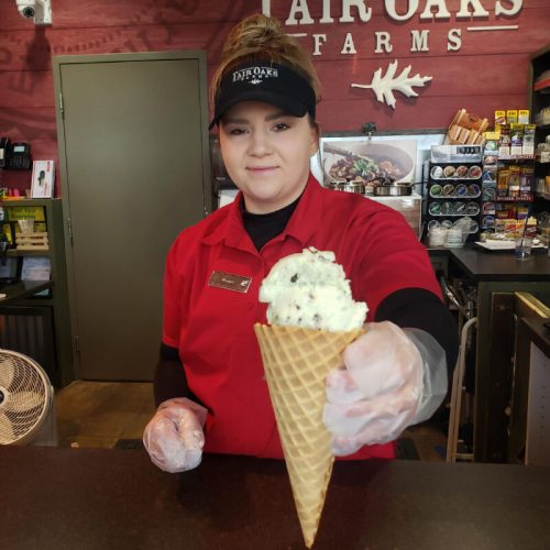 A smiling employee in a red shirt and cap holds out a cone of mint chocolate chip ice cream at a farm stand.