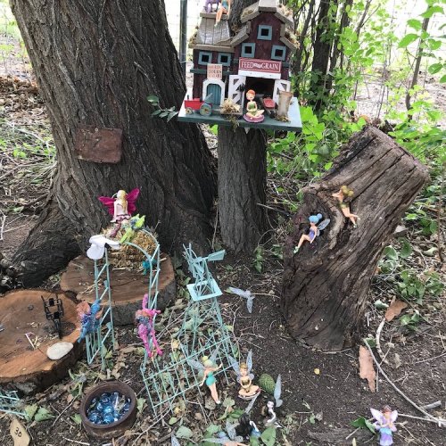 A whimsical fairy garden scene with tiny houses, figures, and decorations nestled among trees and logs.