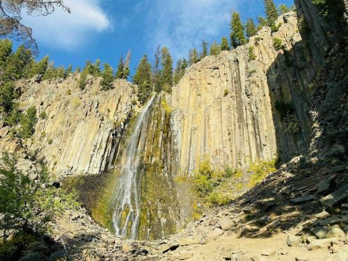 A scenic waterfall cascading down rocky cliffs, surrounded by lush greenery and blue skies.
